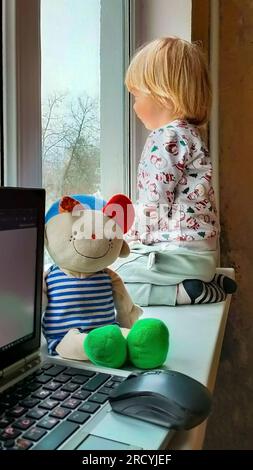 Un petit garçon aux cheveux blonds est assis près de la fenêtre sur le rebord de la fenêtre et regarde par la fenêtre. Hiver. Réveillon du nouvel an. L'enfant est habillé dans une nouvelle année' Banque D'Images