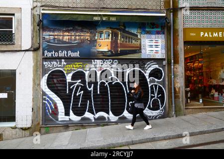 Jeune femme fumant alors qu'elle passe devant des graffitis sur les volets métalliques de la boutique de souvenirs Porto Gift point, Porto / Porto, Portugal Banque D'Images