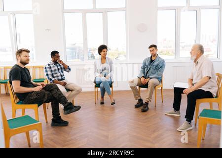 Vue à distance latérale de psychologue coach masculin mature confiant parlant assis en cercle pendant la thérapie de groupe avec des personnes d'âges divers et différents. Banque D'Images
