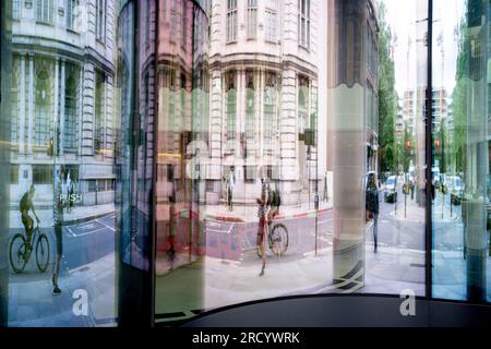 Les travailleurs de la ville et les cyclistes sont reflétés et déformés dans l'entrée vitrée d'un bureau de la City de Londres, le quartier financier de la capitale, le 17 juillet 2023, à Londres, en Angleterre. Banque D'Images