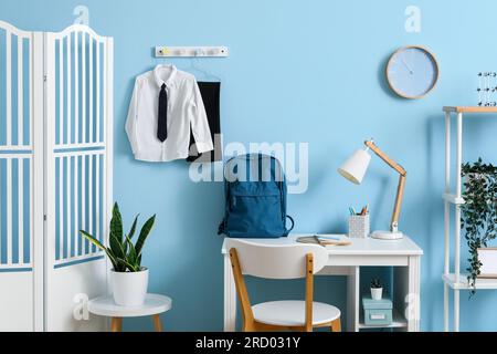 Intérieur du lieu de travail avec bureau, étagère, écran pliant, horloge, sac à dos et uniforme scolaire élégant accroché sur le mur bleu Banque D'Images