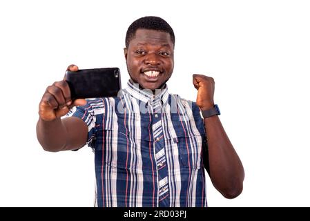un jeune homme en chemise à manches courtes debout sur fond blanc regardant le téléphone portable avec geste de victoire Banque D'Images