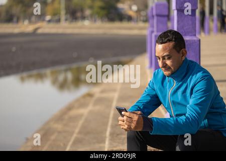 Plan moyen d'un jeune latino en vêtements de sport regardant son téléphone portable dans un parc public. Banque D'Images