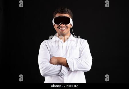 Homme brésilien excité portant des lunettes vision pro vr, debout avec les bras pliés et souriant sur fond de studio noir Banque D'Images