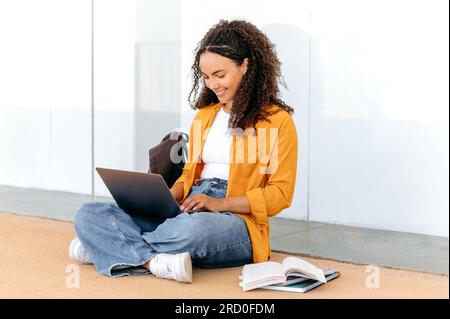 Occupé belle étudiante brésilienne ou hispanique aux cheveux bouclés concentrée et élégante, asseyez-vous près du campus de l'université, en utilisant un ordinateur portable, se préparer à l'examen, faire ses devoirs, tâche de projet, sourire heureux Banque D'Images