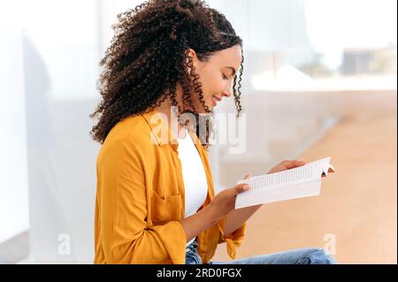 Photo vue de côté d'une belle étudiante brésilienne ou hispanique aux cheveux bouclés élégant, assis près du campus de l'université, concentré à lire son livre préféré, se préparer à l'examen, sourire Banque D'Images
