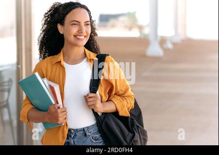 Photo d'une belle étudiante brésilienne ou hispanique aux cheveux bouclés positive, avec un sac à dos, tenir des livres et des cahiers dans sa main, marcher près du campus universitaire, regarder loin et sourire heureux Banque D'Images