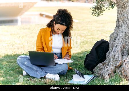 Concept d'apprentissage. Étudiant féminin de race mixte positif moderne, assis sur l'herbe près de l'arbre, avec un ordinateur portable et un bloc-notes, faire ses devoirs, écouter une conférence en ligne, prendre des notes, sourire Banque D'Images