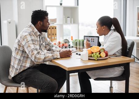 Vue d'épaule d'une femme afro-américaine analysant les données nutritionnelles d'un patient sur tablette numérique en salle de consultation. Diététiste efficace évaluant les besoins de santé du client masculin dans un centre médical. Banque D'Images