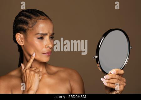 Femme noire frustrée d'âge moyen tenant le miroir et touchant la joue, souffrant de problèmes de peau ou remarquant des changements d'âge Banque D'Images