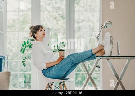 Ordinateur de bureau femme. Elle est vêtue d'une chemise blanche et d'un Jean bleu, s'assoit à une table sur laquelle il y a un ordinateur portable et un bloc-notes, met ses pieds sur le Banque D'Images