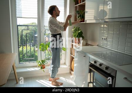 Homme portant corset de colonne vertébrale pour fixer le bas du dos pulvérisant des plantes sur la cuisine à la maison en congé de maladie. Banque D'Images