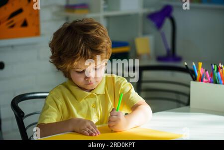 Petit écolier faisant ses devoirs assis à son bureau. Enfant garçon de l'école primaire. Petit écolier avec des crayons colorés. Dessins enfants. Créativité Banque D'Images