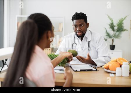 Homme afro-américain concentré dans le manteau blanc tenant l'avocat tout en parlant avec la cliente dans la salle de conseil. Médecin spécialiste de l'alimentation encourageant à enrichir manger avec des fruits pleins de nutriments. Banque D'Images