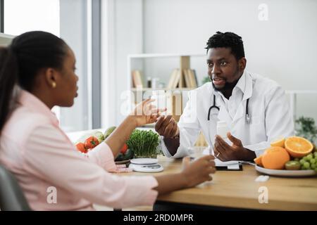 Bel homme afro-américain en manteau blanc donnant des pilules bouteille à charmante femme dans la salle de consultation de l'hôpital. Professionnel de la nutrition masculine améliorant la santé globale avec des compléments alimentaires. Banque D'Images