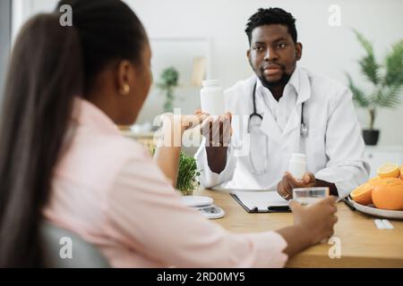 Bel homme afro-américain en manteau blanc donnant des pilules bouteille à charmante femme dans la salle de consultation de l'hôpital. Professionnel de la nutrition masculine améliorant la santé globale avec des compléments alimentaires. Banque D'Images