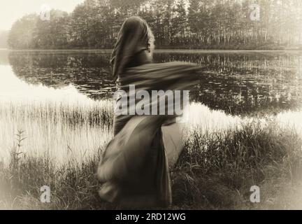 Chaman femme en robe à capuche, dansant dans un mouvement flou. Elle salue le matin d'un lac magique. La photo évoque une qualité fantôme d'un autre monde. Banque D'Images