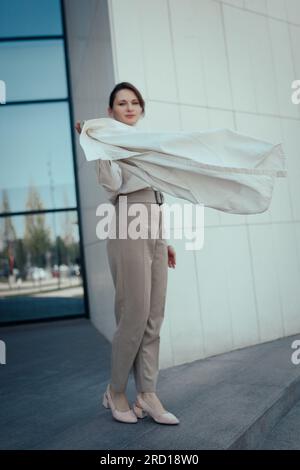 Portrait d'une femme d'affaires mignonne réussie dans un costume pantalon beige élégant debout près de l'immeuble de bureaux, vertical Banque D'Images