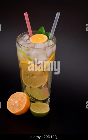 Limonade rafraîchissante dans un grand verre à facettes avec de la glace, des pailles et des morceaux d'agrumes sur fond noir, à côté de tranches d'orange et de citron. Verti Banque D'Images