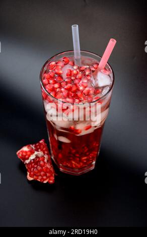 Un grand verre de jus de grenade avec de la glace, des graines et des tubes sur un fond noir, à côté se trouve un fruit mûr cassé. Gros plan. Banque D'Images