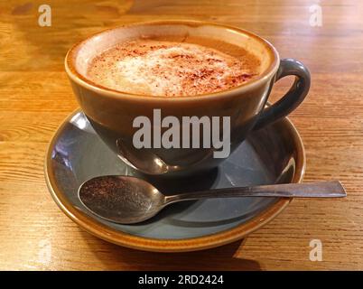 Café cappuccino servi dans une tasse en céramique avec soucoupe et cuillère sur une table en bois, photographié dans un cadre de café communautaire Banque D'Images