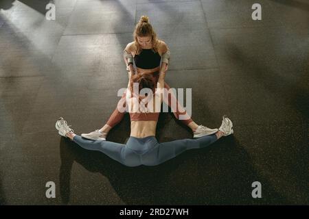 Vue de dessus de l'entraîneur aidant la femme à s'asseoir sur straddle split sur le sol dans la salle de gym Banque D'Images
