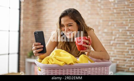 Jeune belle femme hispanique utilisant un smartphone buvant du café penché sur le panier avec des vêtements à la buanderie Banque D'Images