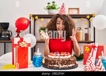 Femme hispanique avec les cheveux bouclés célébrant l'anniversaire tenant le grand gâteau au chocolat célébrant la victoire avec le sourire heureux et l'expression du gagnant avec Raised Banque D'Images