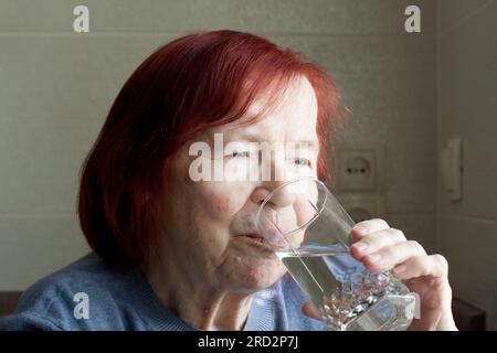 femme âgée buvant de l'eau minérale du verre. Portrait en gros plan de femme senior aux cheveux rouges en pull bleu, apaise la soif, à la maison. De l'eau pour de la santé Banque D'Images