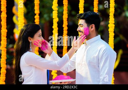 Heureux jeune couple indien appliquant des couleurs de Holi sur le visage ensemble en tenant le fond de décoration de fleur de plaque de puissance de Holi - concept de festival de Holi Banque D'Images