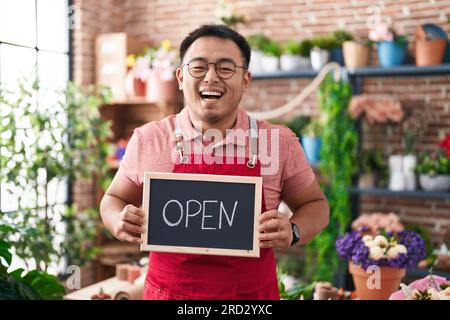 Jeune homme chinois travaillant au fleuriste tenant un signe ouvert souriant et riant fort à haute voix parce que drôle de blague folle. Banque D'Images