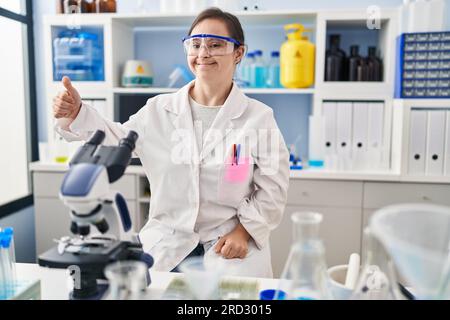 Fille hispanique avec le syndrome de Down travaillant au laboratoire scientifique semblant fière, souriante faisant le geste pouce vers le haut sur le côté Banque D'Images