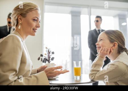 service de sécurité, concept de sécurité privée, femme blonde joyeuse avec tasse de café parlant avec la fille preteen dans le café, gardes du corps sur le backgr flou Banque D'Images