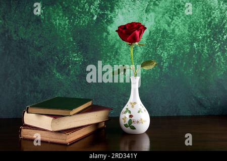 Vase fleur en céramique avec rose rouge unique et vieux livres sur un dessus de table poli avec fond tacheté Banque D'Images