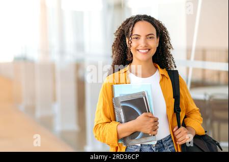 Heureuse belle étudiante brésilienne ou hispanique aux cheveux bouclés, avec un sac à dos, tenir des livres et des cahiers dans sa main, se tenir près du campus universitaire, regarder et sourire à la caméra. Copy-space Banque D'Images