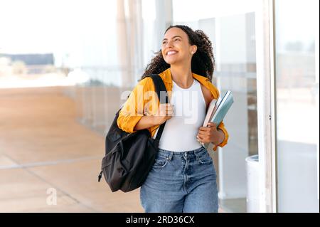 Heureuse belle étudiante brésilienne ou hispanique aux cheveux bouclés, avec un sac à dos, tenir des livres et des cahiers dans sa main, marcher près du campus universitaire, regarder loin et sourire, a terminé la journée d'école Banque D'Images