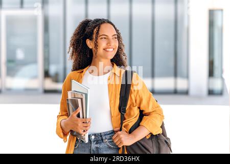 Heureuse étudiante. Étudiante positive, de nationalité brésilienne ou hispanique, avec un sac à dos, tenant des livres et des cahiers dans sa main, se tenir près du campus universitaire, regarde loin et souriant Banque D'Images
