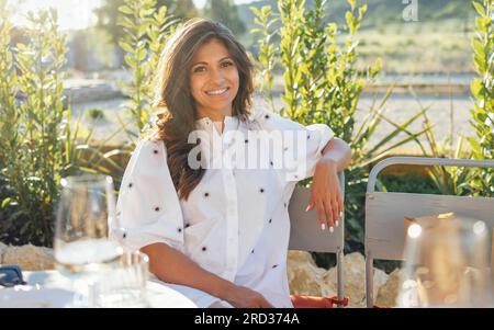 Jeune belle brunette est relaxant dans un café en plein air. Une charmante fille dans des vêtements élégants sourit et s'assoit à une table dans un restaurant. Verres à vin Banque D'Images