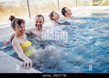 Deux couples mariés discutent et profitent d'une piscine extérieure. Les amis rient et se détendent dans l'eau tourbillonnante. Les jeunes se détendent dans un spa ou un hôtel. Banque D'Images