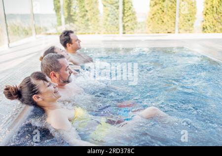 Deux couples mariés discutent et profitent d'une piscine extérieure. Les amis rient et se détendent dans l'eau tourbillonnante. Les jeunes se détendent dans un spa ou un hôtel. Banque D'Images