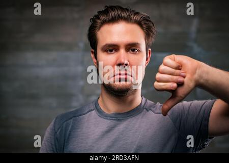 Jeune bel homme pouce vers le bas portrait sur fond de mur gris Banque D'Images