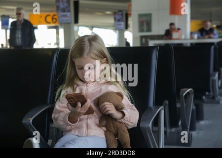 Petite fille assise dans un hall de départ d'aéroport tenant un ours en peluche et jouant avec plaisir sur son téléphone portable Banque D'Images