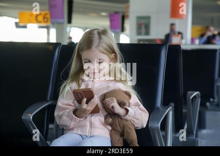 Petite fille assise dans un hall de départ d'aéroport tenant un ours en peluche et jouant avec plaisir sur son téléphone portable Banque D'Images