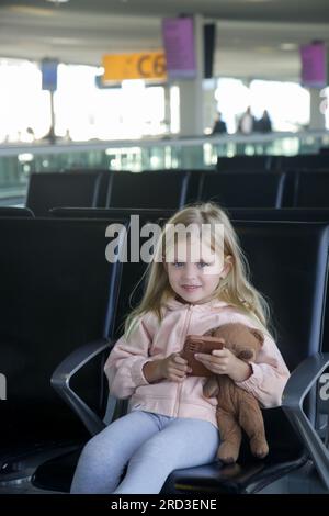 Petite fille assise dans un hall de départ d'aéroport tenant un ours en peluche et jouant avec plaisir sur son téléphone portable Banque D'Images