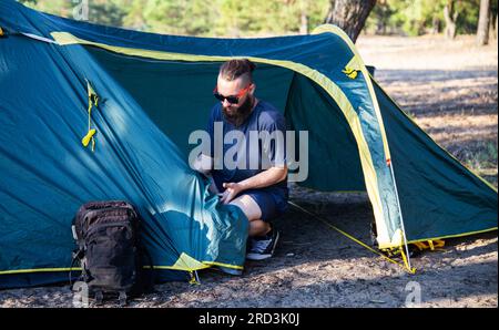 Un beau type avec une barbe et des lunettes noires installe une tente touristique sur la rive sablonneuse de la rivière dans la forêt. Activités de plein air en été. Mannequin Banque D'Images