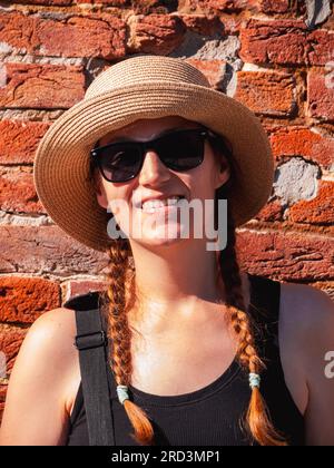 Jeune femme dans le portrait de chapeau de paille sur fond urbain de mur de brique rouge. Fille millénaire marchant sur la rue de la ville d'été avec le soleil lumineux. Conf. Élégante Banque D'Images