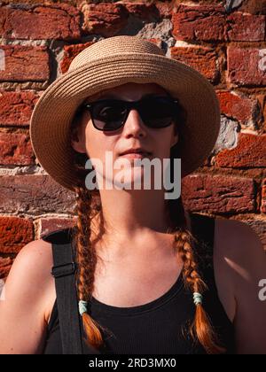 Jeune femme dans le portrait de chapeau de paille sur fond urbain de mur de brique rouge. Fille millénaire marchant sur la rue de la ville d'été avec le soleil lumineux. Conf. Élégante Banque D'Images