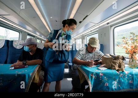 (230718) -- MENGLA, 18 juillet 2023 (Xinhua) -- Yi Bofeng (C) sert les passagers à bord du train D887 le 16 juillet 2023. Yi Bofeng est originaire du comté de Mengla dans la province du Yunnan. Après avoir obtenu son diplôme de major laotien dans un collège technique de la capitale provinciale Kunming en 2020, elle a été recrutée comme hôtesse de train dans China Railway Kunming Bureau Group Co., Ltd Le chemin de fer Chine-Laos, un projet historique de coopération de haute qualité ceinture et route, a commencé ses opérations en décembre 2021. Yi était à bord du premier train du chemin de fer, témoin du moment historique de l'exploitation ferroviaire. Le 1 035 Banque D'Images