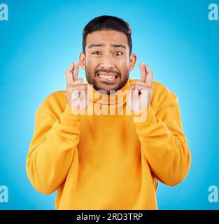 Nerveux, portrait et doigts croisés par l'homme asiatique avec anxiété en studio pour des nouvelles, des commentaires ou un examen sur fond bleu. Main, emoji et visage de gars Banque D'Images