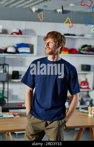 Jeune designer rousse dans des vêtements décontractés tenant les mains dans les poches et debout près de la table de travail dans le studio d'impression floue, l'entrepreneuriat pratique c Banque D'Images